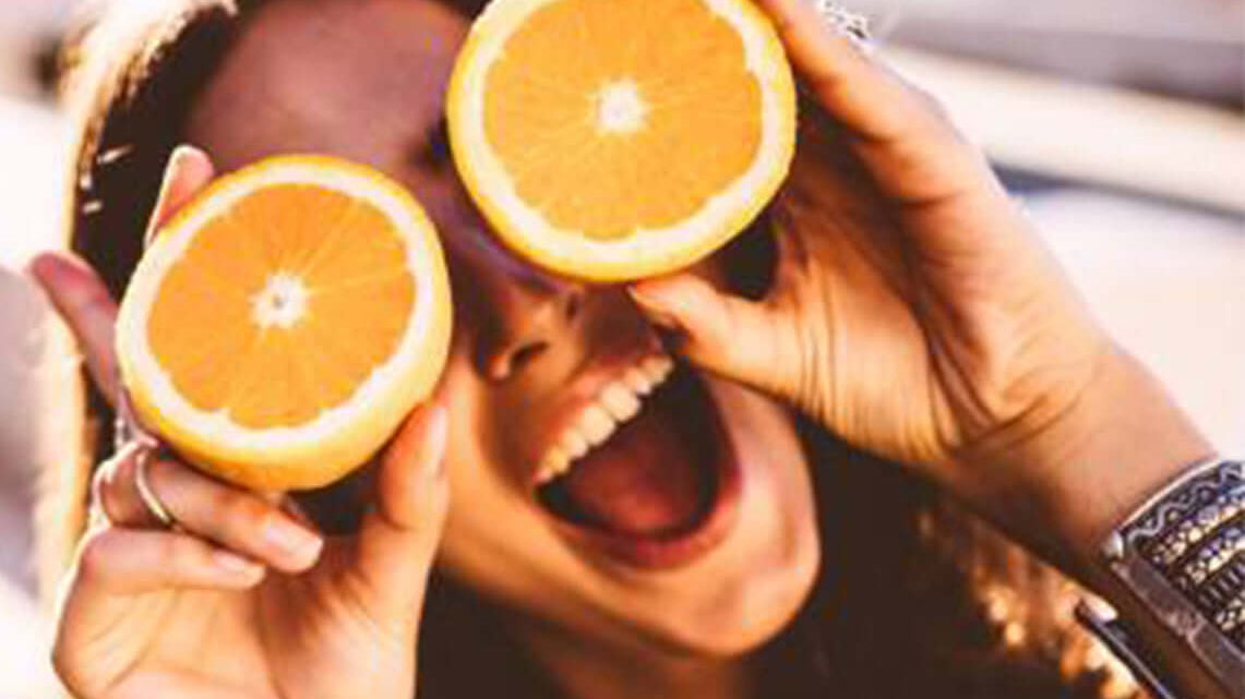 Young woman holding two orange slices over her eyes laughing as she looks up