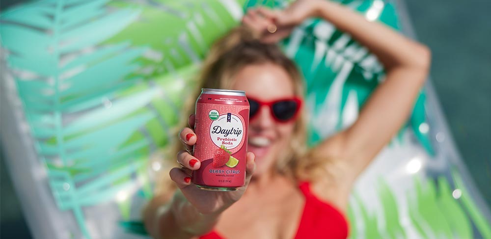 A woman lying on a raft in a pool in sunglasses holding up a can of Daytrip prebiotic soda.
