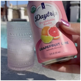 A woman holding a can of Daytrip grapefruit lime prebiotic soda in front of a glass with ice at a swimming pool