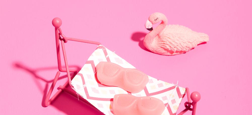 Miniature photo scene showing two sunglass-shaped pink gummies lying on a tiny hammock on a pink background, with a flamingo resting next to it.