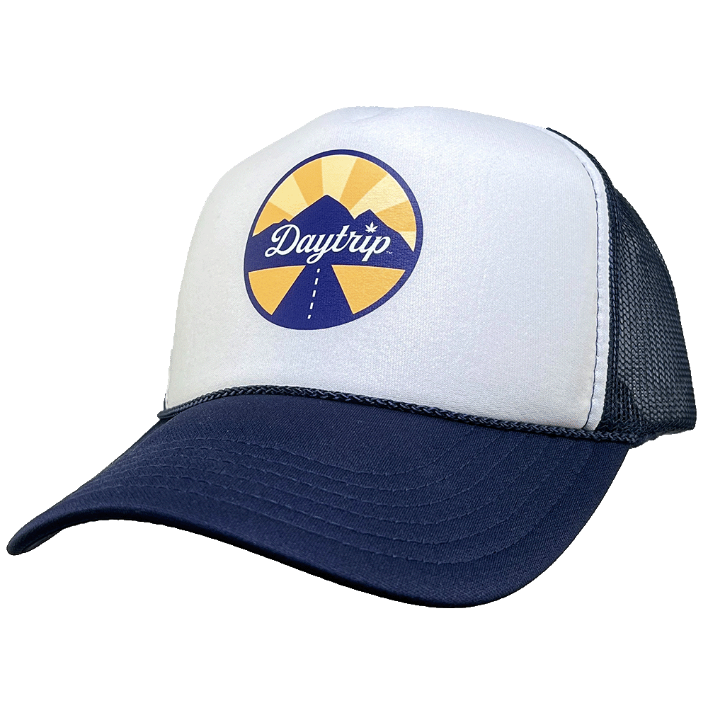 Front view of the Daytrip mountain trucker hat. Navy blue brim and mesh back, with a white front. On the white front is a circular lockup featuring the Daytrip script logo laid over top of a blue and yellow mountain scene, with a highway vanishing in the