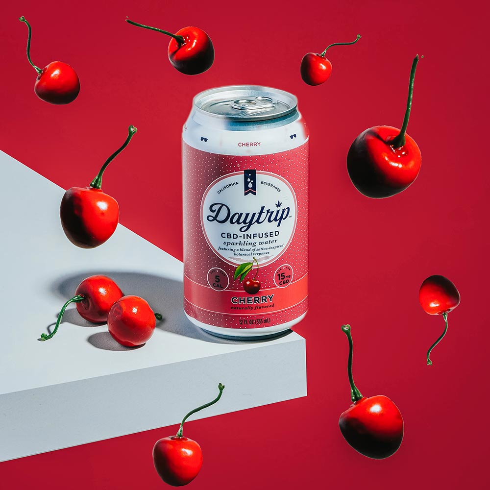 A photorealistic render of a can of Daytrip cherry sparkling water sitting on the corner of a white countertop, with a deep red background and cherries floating in the space around the can.