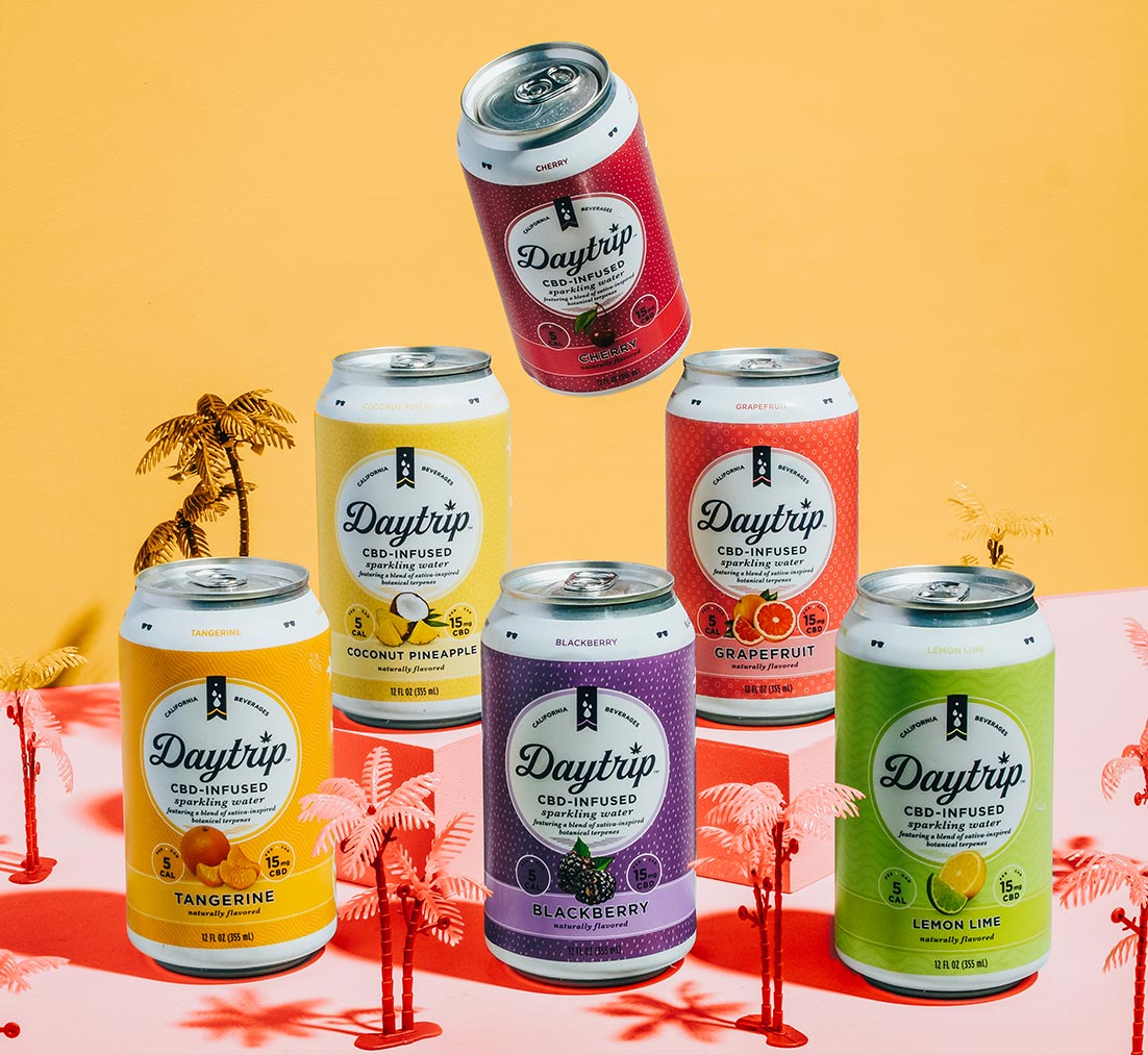 A miniature lifestyle scene showing all six flavors of Daytrip CBD infused sparkling water stacked in tiers with miniature palm trees scattered between. One can floats above the top two tiers and is angled like it's about to pour.