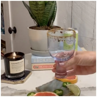 A person holding a cocktail glass with flavored salt on the rim, and cut up citrus fruit and jalapeños in the foreground with a candle, plant and books in the background.
