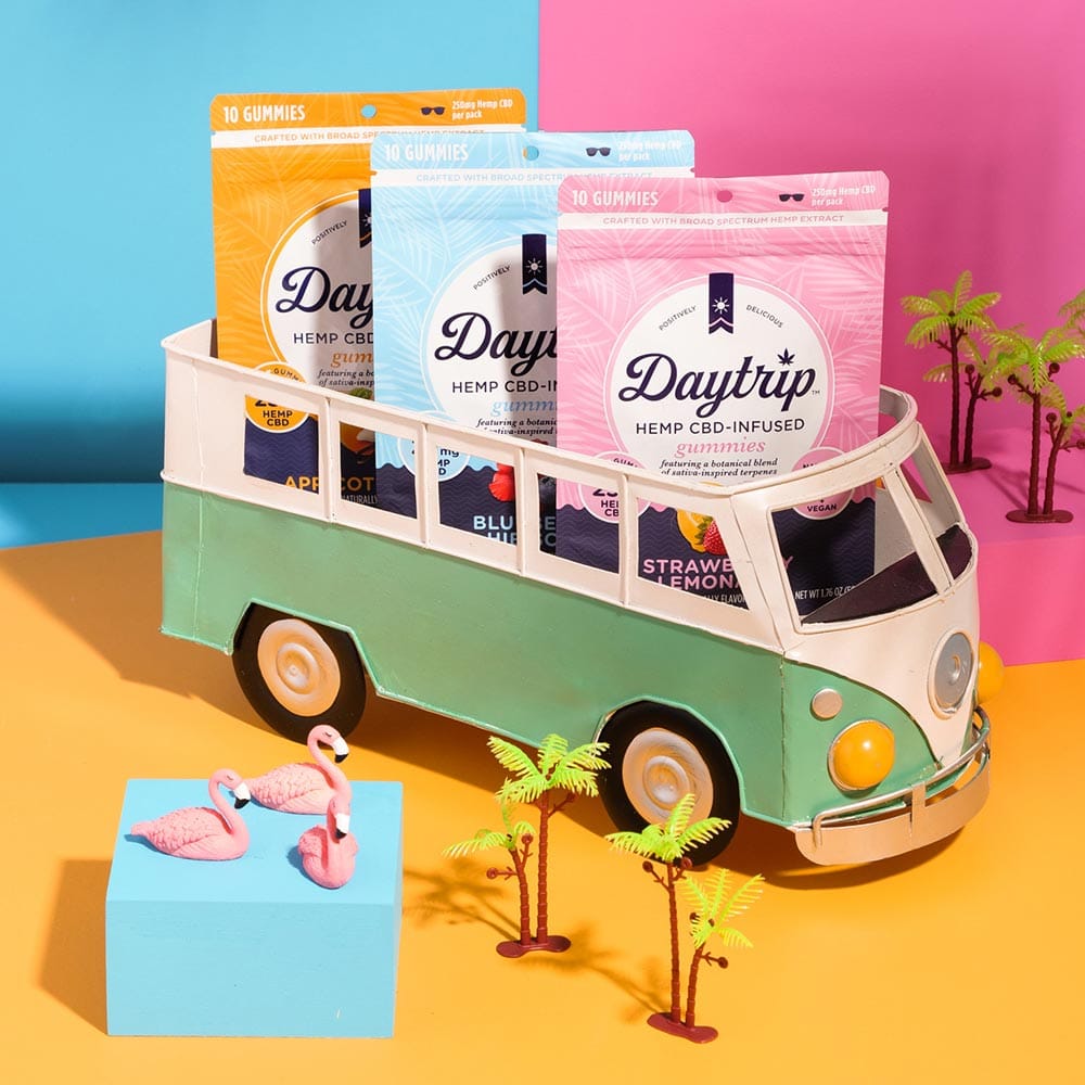 A miniature lifestyle scene featuring bags of Daytrip gummies in a green VW van sitting on an orange surface. Next to the van are miniature palm trees and miniature flamingos sitting on a stack of blue post-it notes. More palm trees on a pink surface sit in the background.