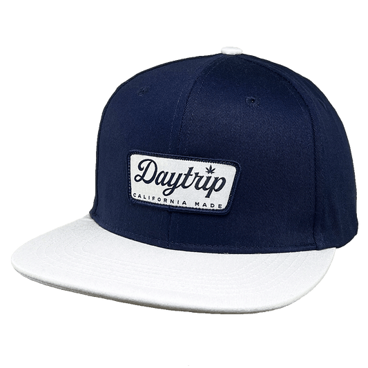 California Snapback hat on a white background. Navy blue / denim upper hat with a white "california made" Daytrip logo patch with blue embroidered stitching. White brim on hat.