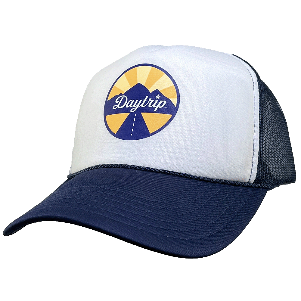 Front view of the Daytrip mountain trucker hat. Navy blue brim and mesh back, with a white front. On the white front is a circular lockup featuring the Daytrip script logo laid over top of a blue and yellow mountain scene, with a highway vanishing in the 