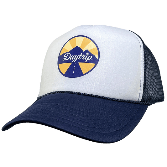 Front view of the Daytrip mountain trucker hat. Navy blue brim and mesh back, with a white front. On the white front is a circular lockup featuring the Daytrip script logo laid over top of a blue and yellow mountain scene, with a highway vanishing in the 